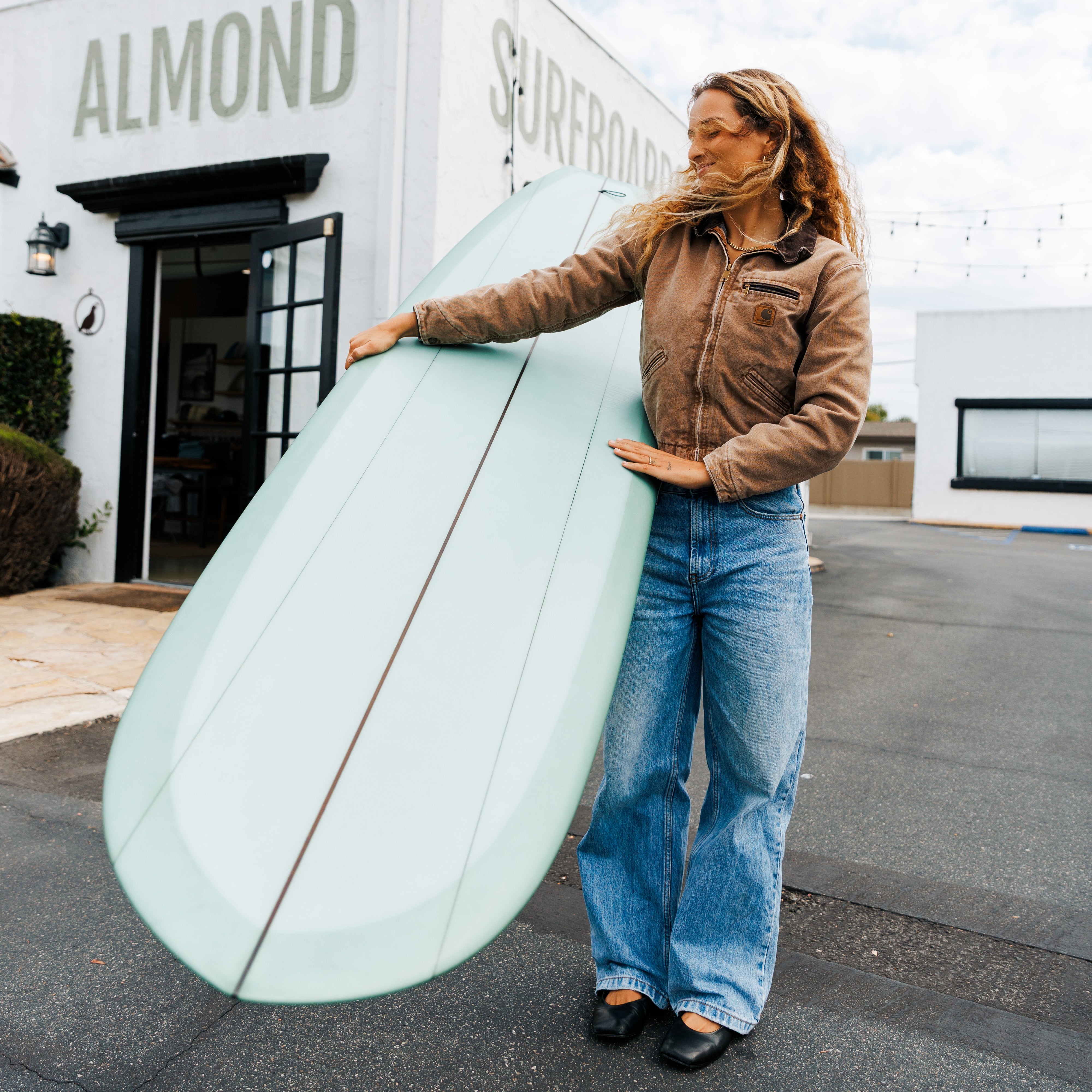 Person holding a surfboard in front of a building with 'Almond Surfboards' on it.